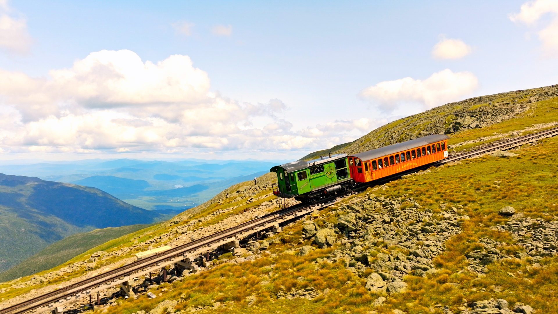 a green and red train high on a mountain top with views of mountains in the background.