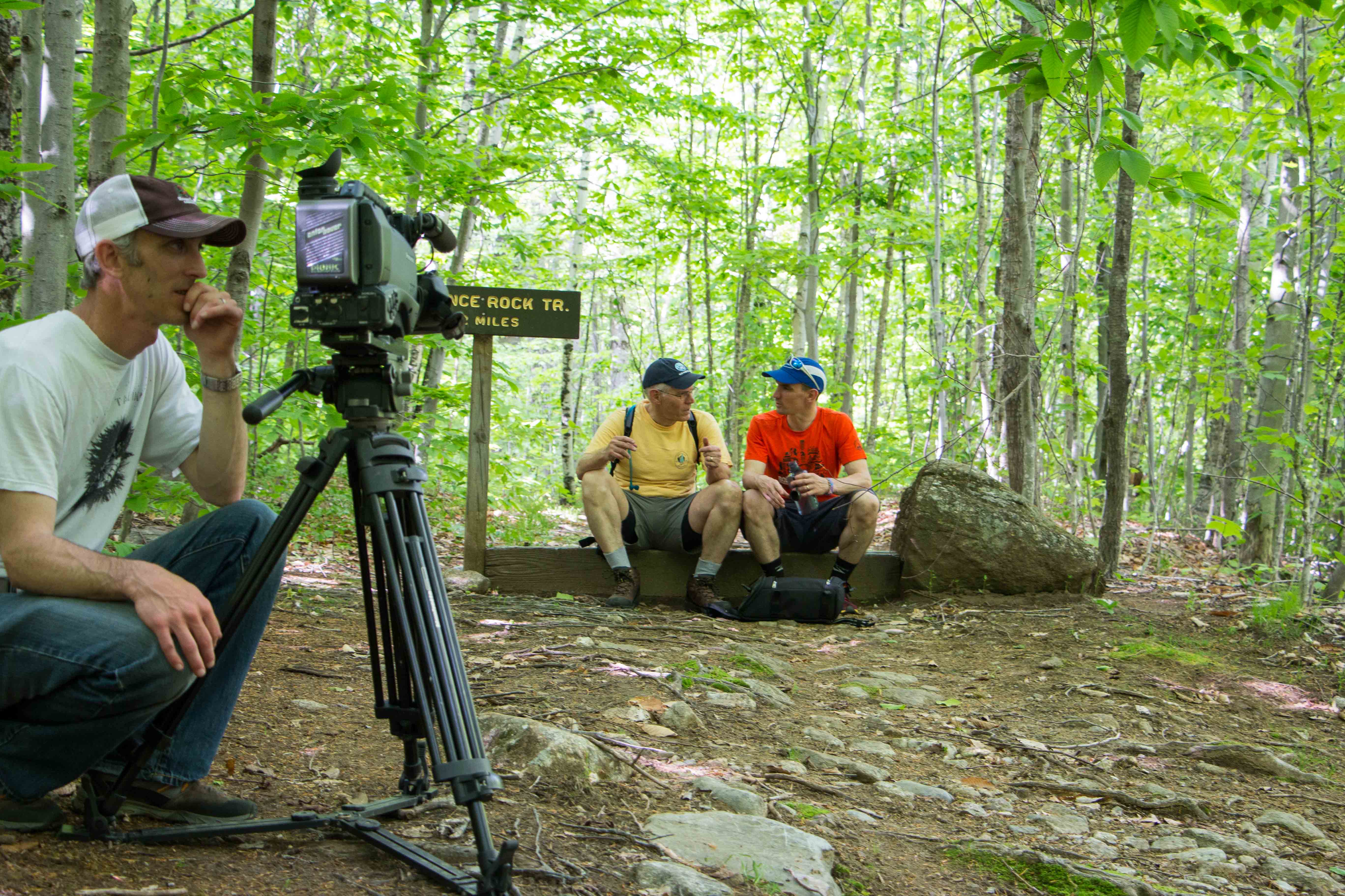 a man with a camera pointed at two hikers sitting on a rock