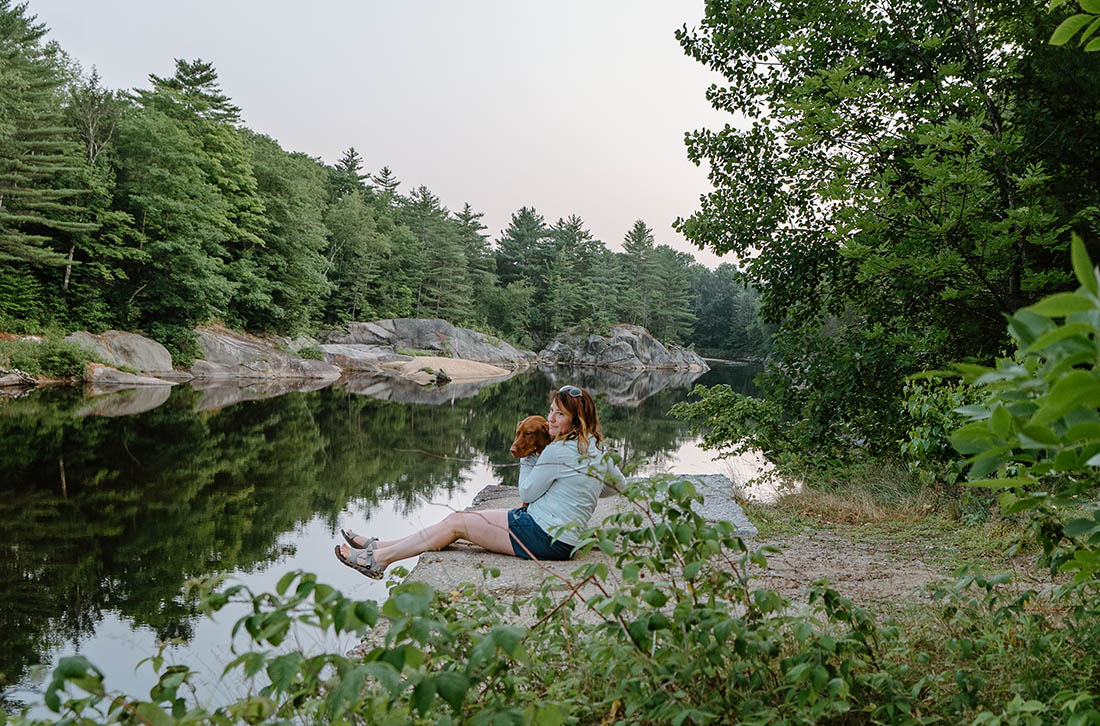 a woman and her dog sitting on a rock at a river